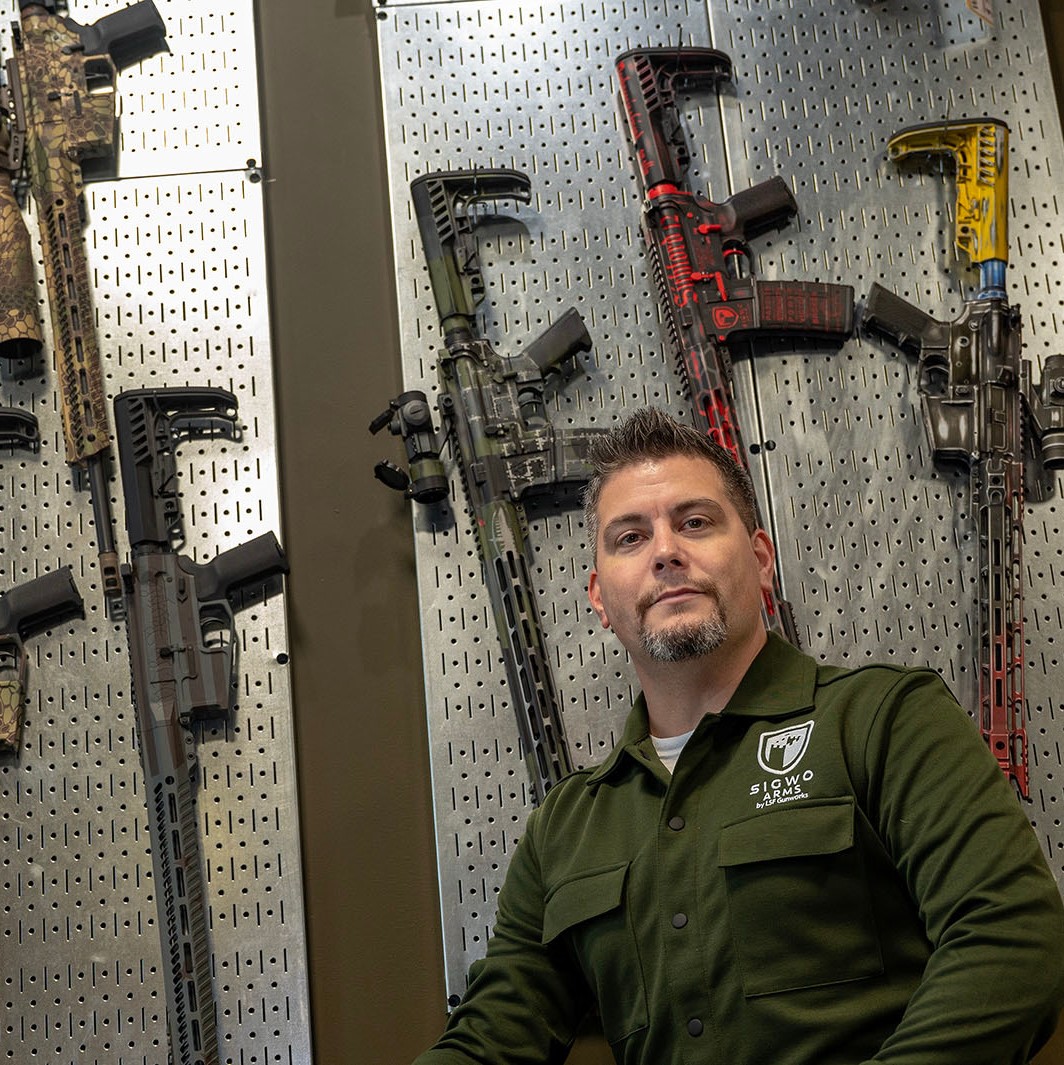 A man poses for a photo in front of guns displayed on a wall