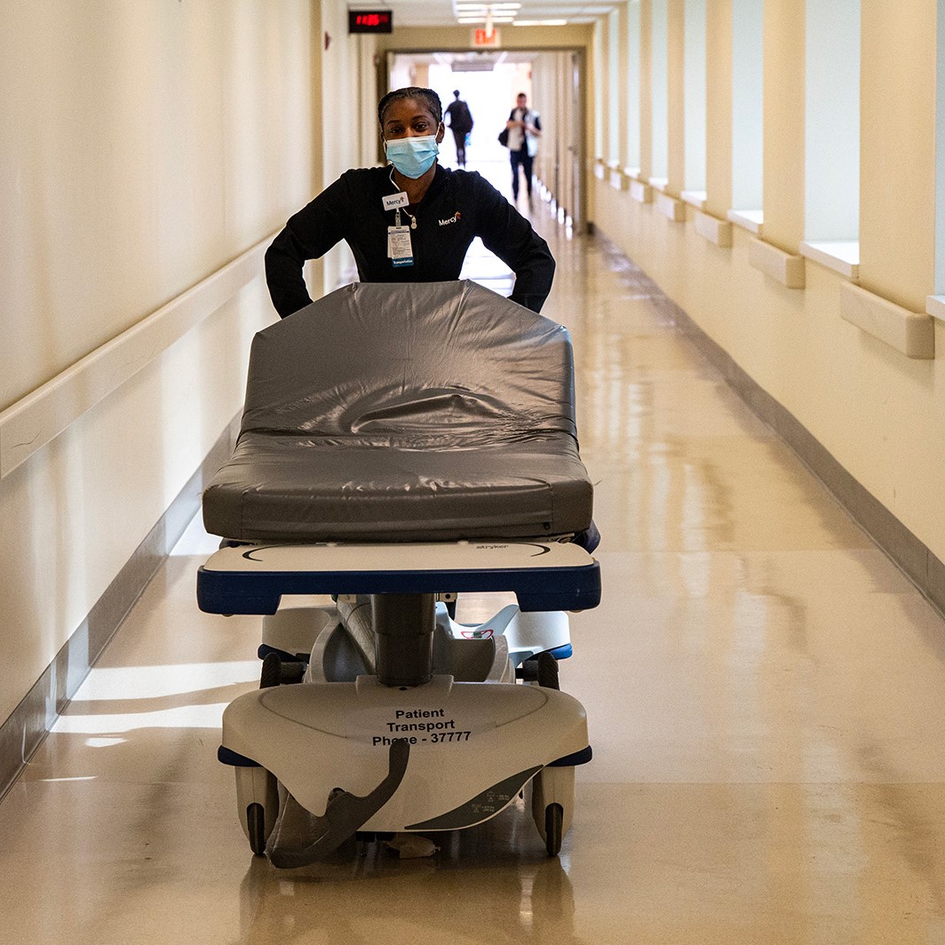 A hospital worker pushes a patient transporter down a long hallway