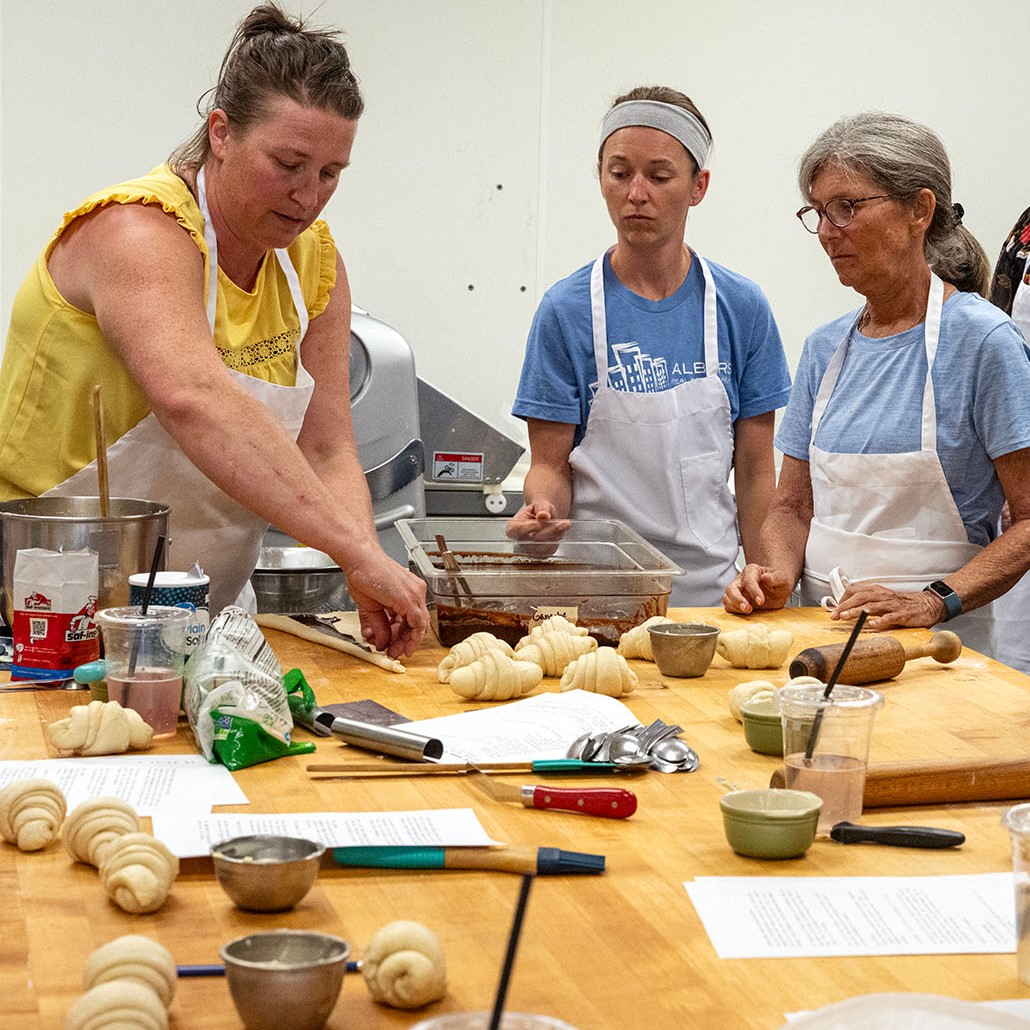 Two women watch as the instructor in a cooking class demonstrates how to make croissants
