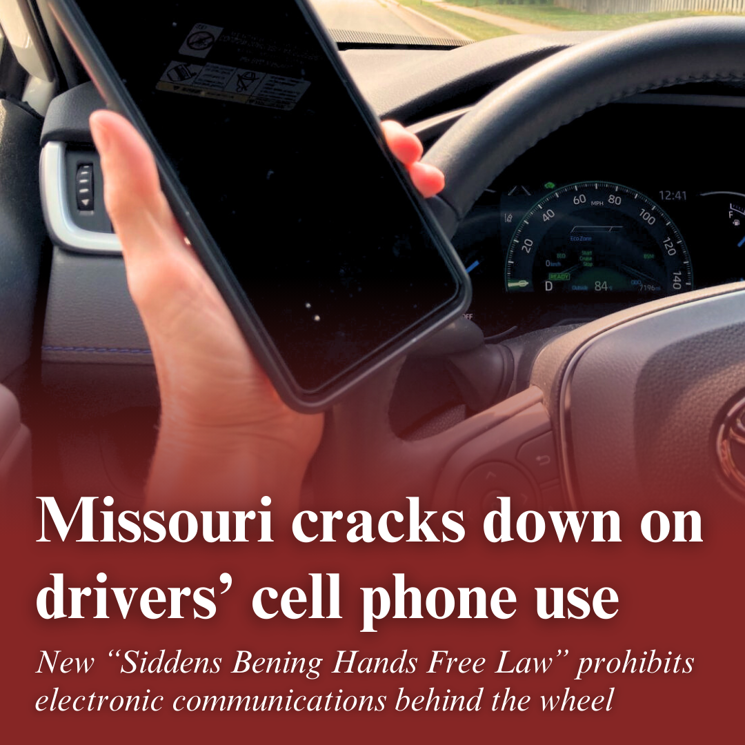 A hand holds a cell phone next to a car's steering wheel