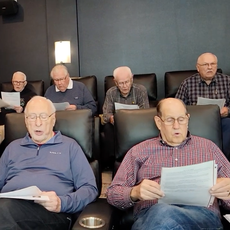 A group of men, seated in chairs, sing a hymn