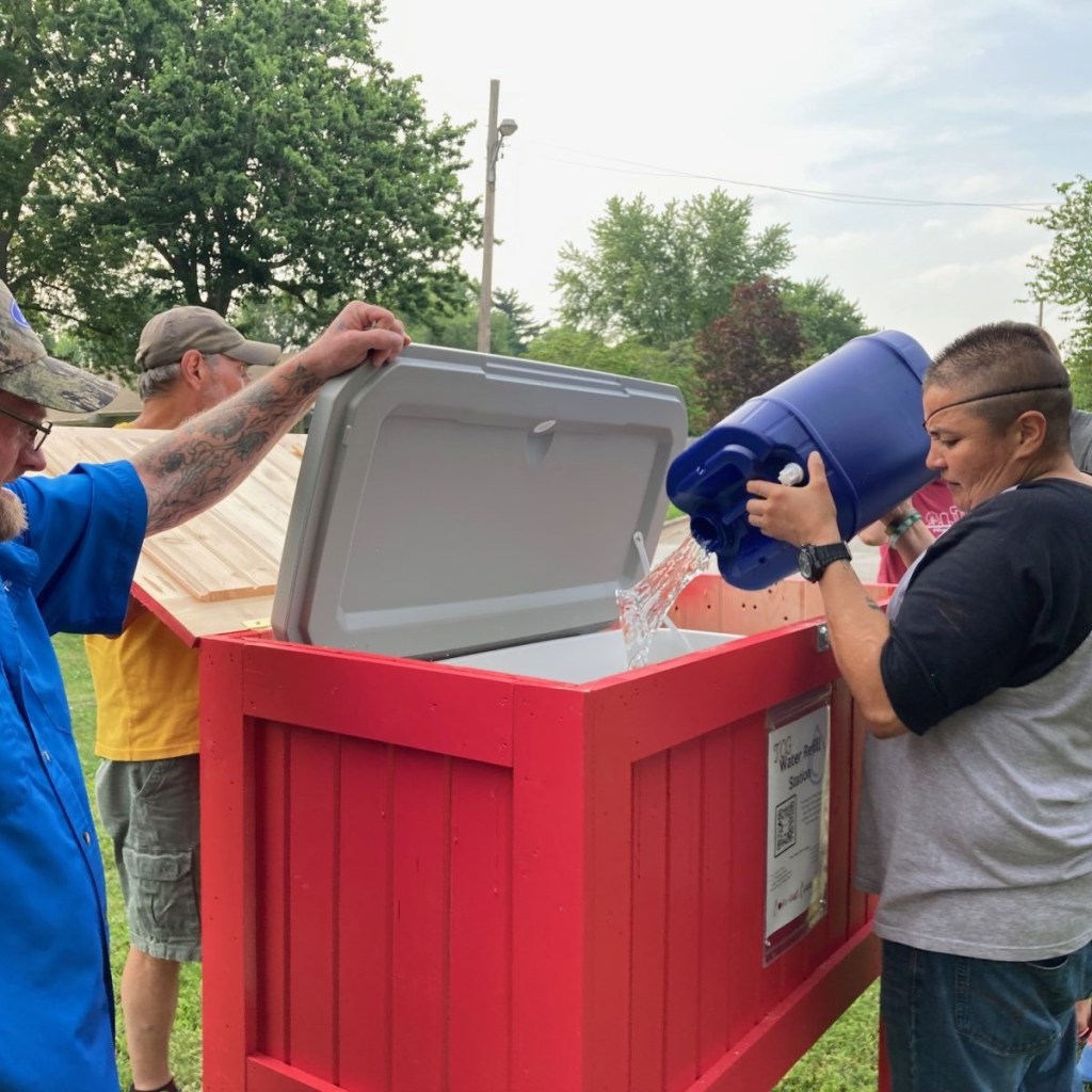 Connecting Grounds Lived Experience Cohort member T.J. Potter holds the lid while Alex (left) dumps water into a refill station near National Avenue Christian Church.