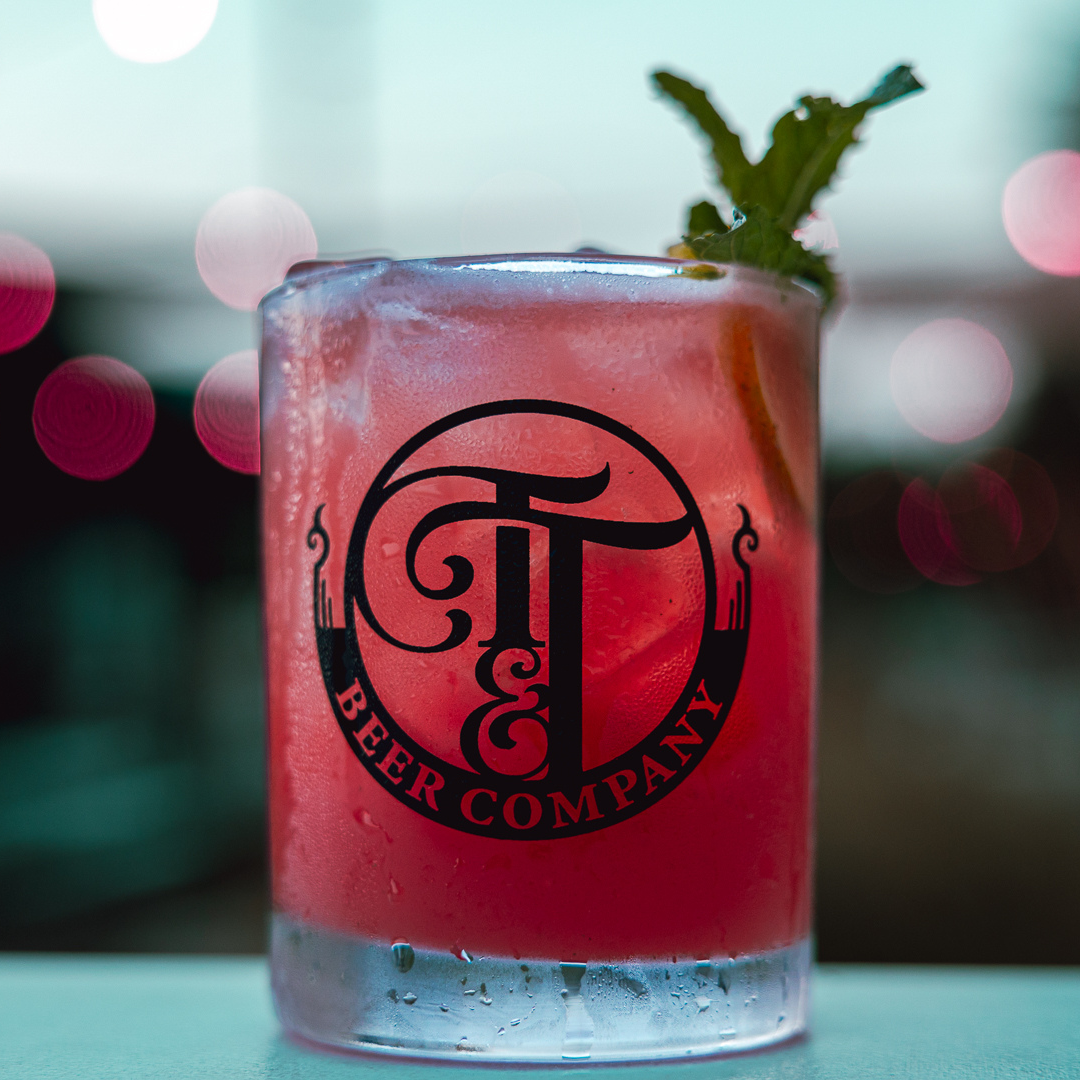 A purple-hued mocktail in a glass with the Tie & Timber Beer Logo printed on the side of it