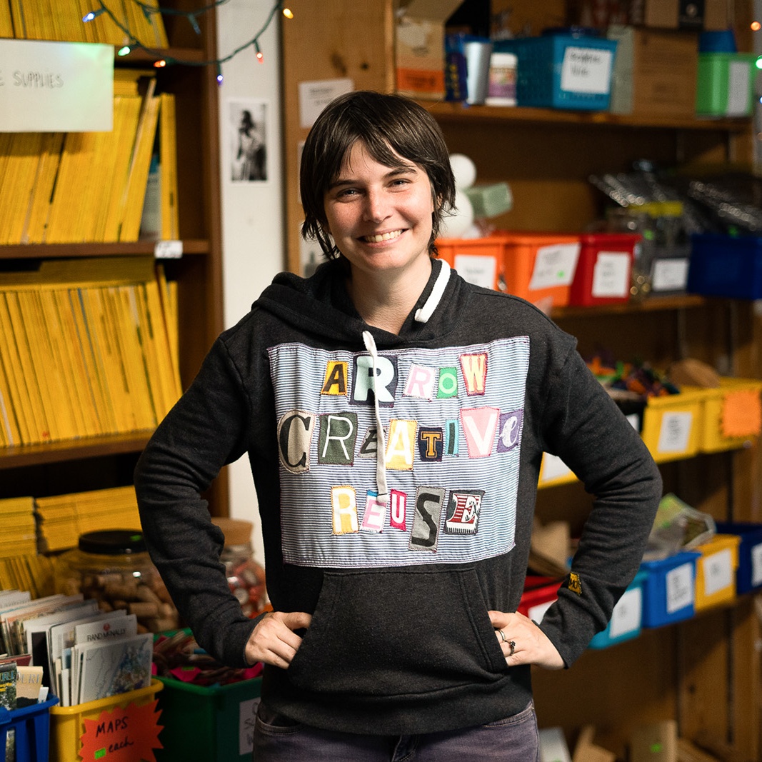 A woman in a black sweatshirt reading "Arrow Creative Reuse" stands in front of shelves of art supplies
