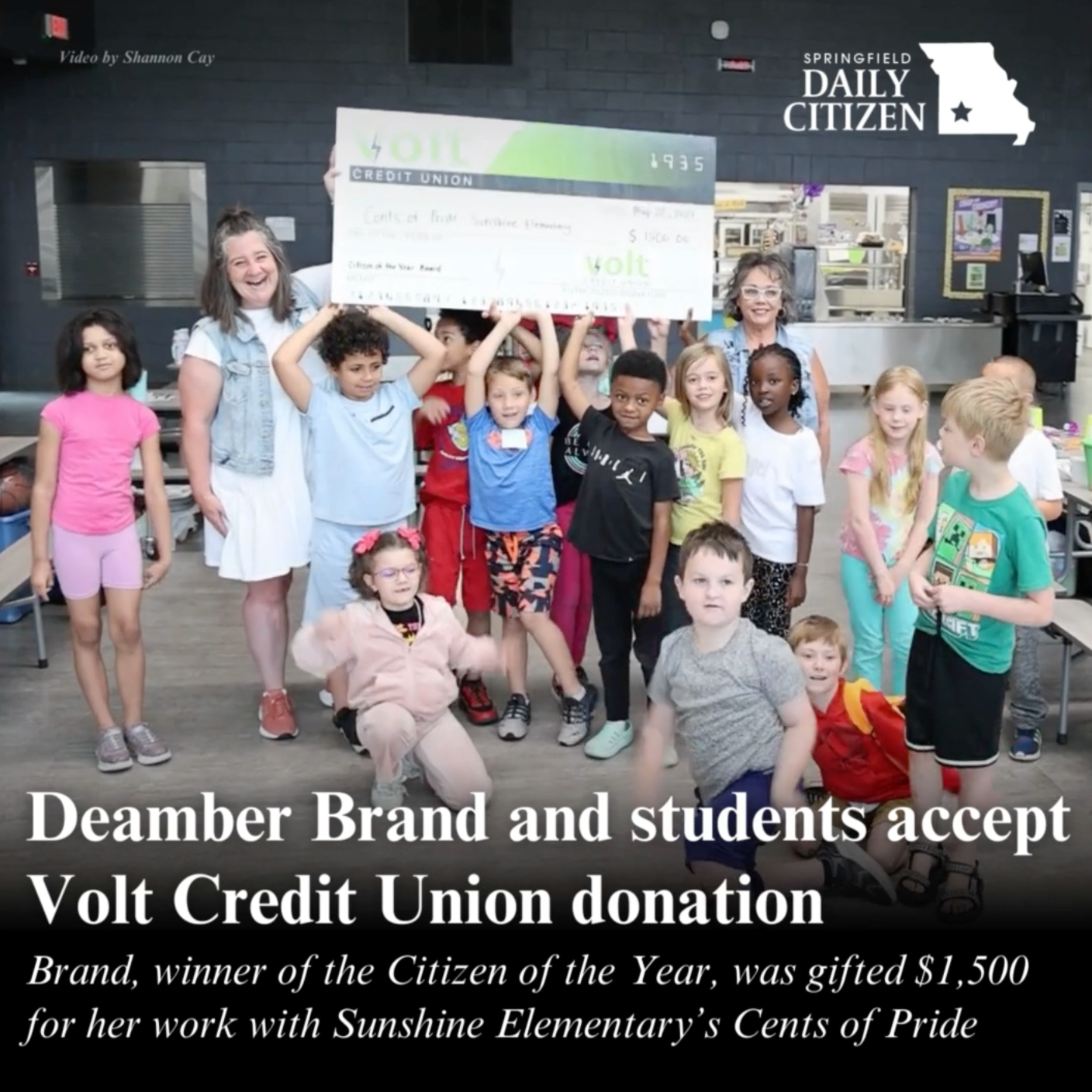 Deamber Brand and several Sunshine Elementary students hold up a giant check in the school's cafeteria. Text on the image reads: "Deamber Brand and students accept Volt Credit Union donation. Brand, winner of Citizen of the Year, was gifted $1,500 for her work with Sunshine Elementary's Cents of Pride."