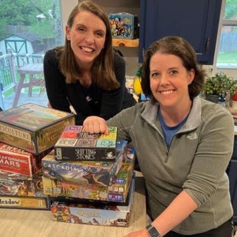 Two women pose with a stack of board game boxes