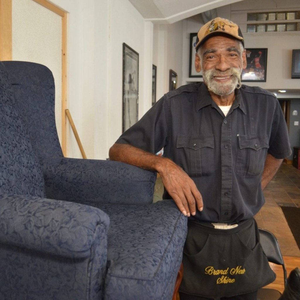 Larry Hairston stands next to his shoeshine chair. He's wearing a hat and a black apron that reads "Brand New Shine"