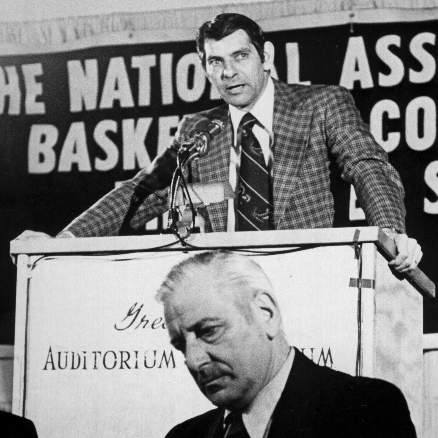 A man in a suit stands behind a podium, giving a speech