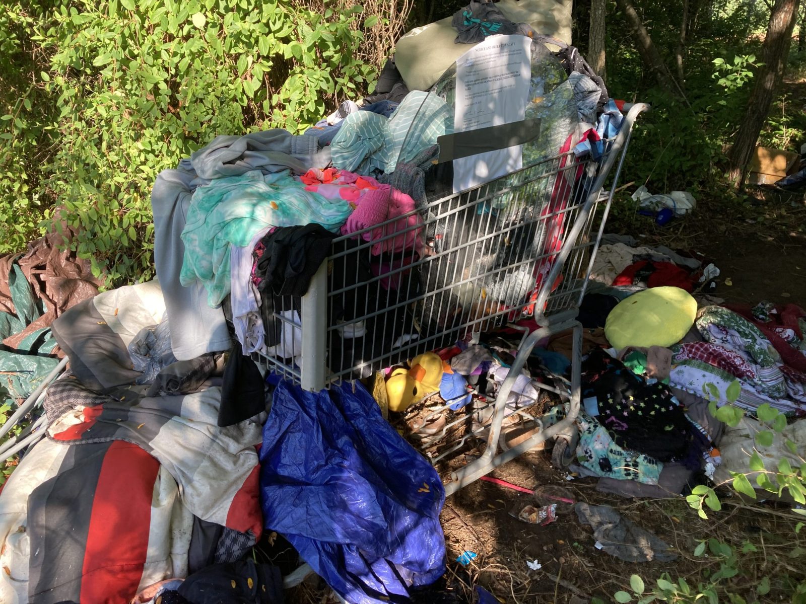 This is a photo of a shopping cart filled with clothing and blankets at the site of a former homeless camp. A trespassing notice has been attached to the shopping cart.