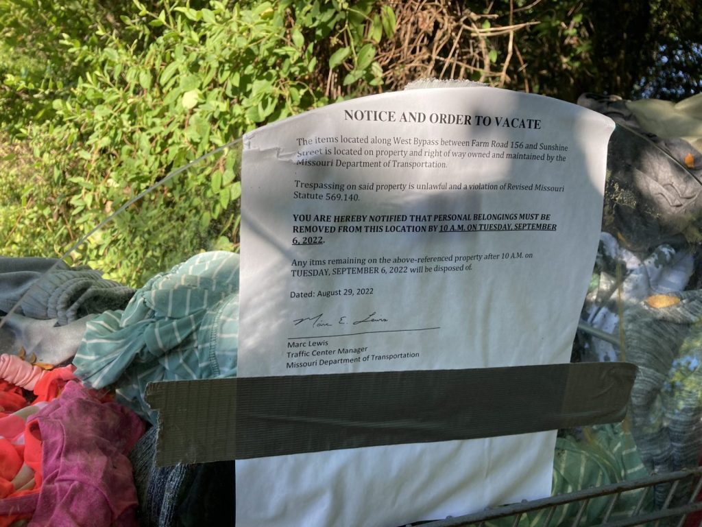 This is a notice and order to vacate attached to a shopping cart filled with clothes. The shopping cart is sitting at the edge of the woods where homeless people were camping.
