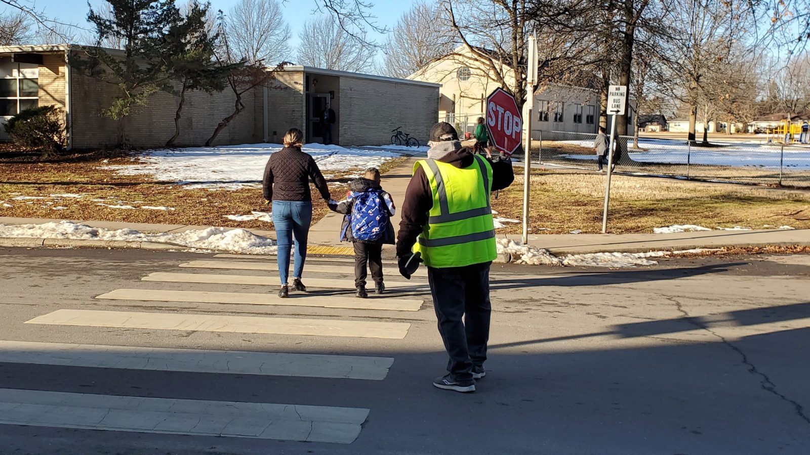 A mother escorts her child across a street as a crossing guard holds a stop sign
