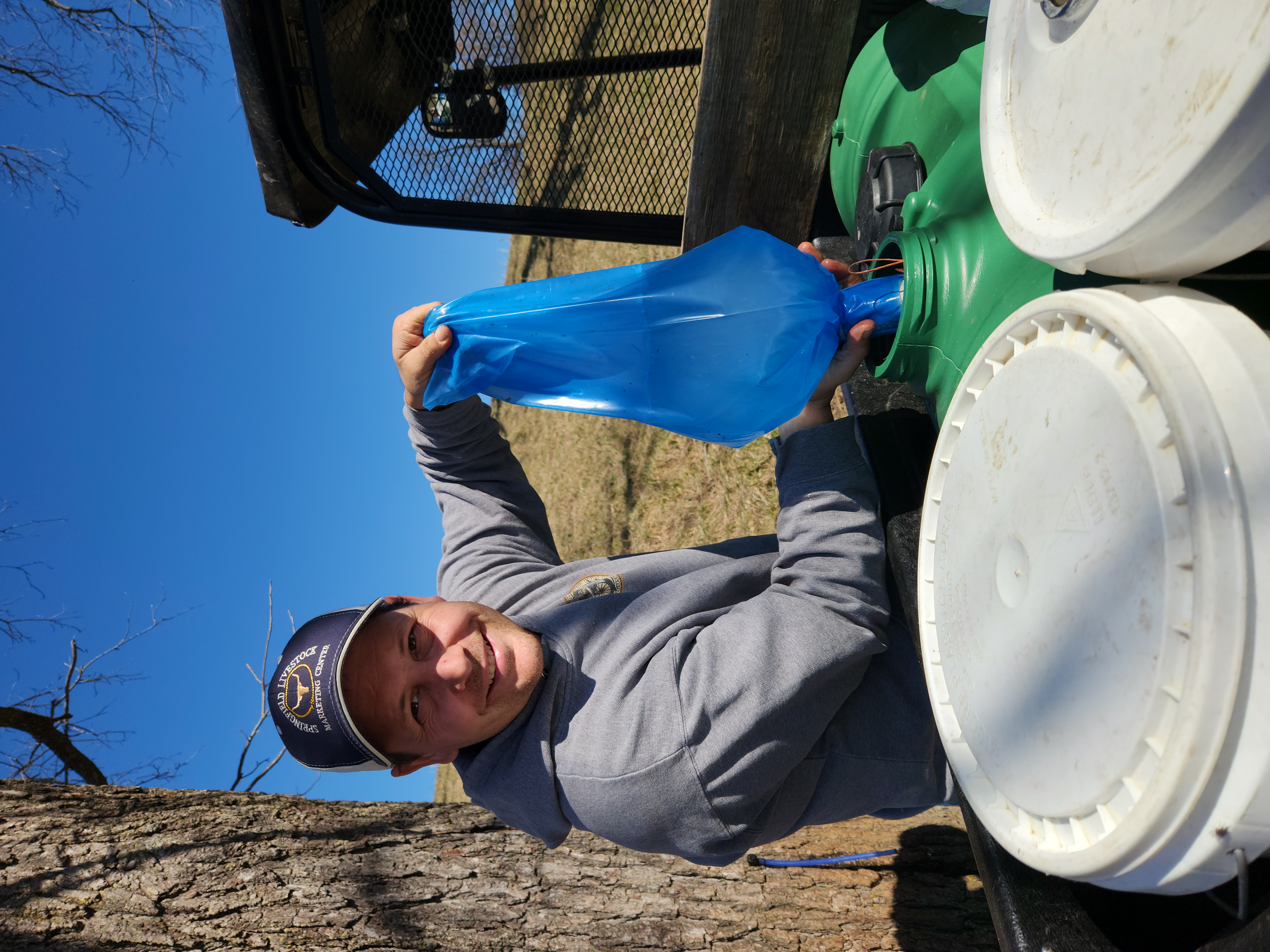 A smiling man pours tree sap from a blue bag into a green plastic container