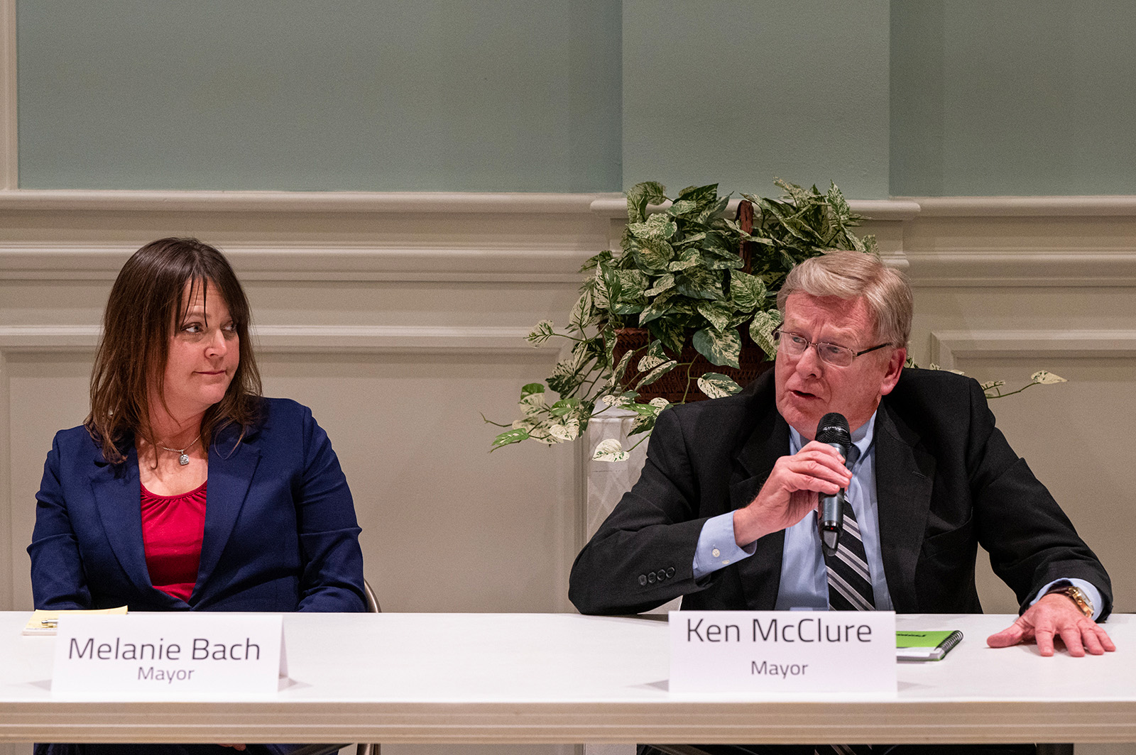 Two candidates for mayor, a man and a woman, sit side-by-side at a community forum