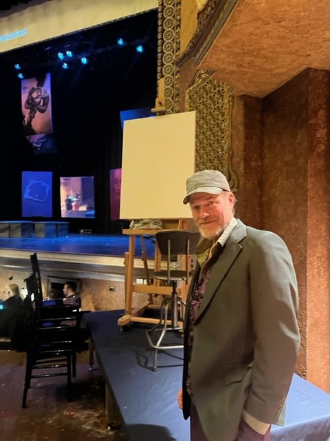 An artist stands next to a blank canvas on an easel