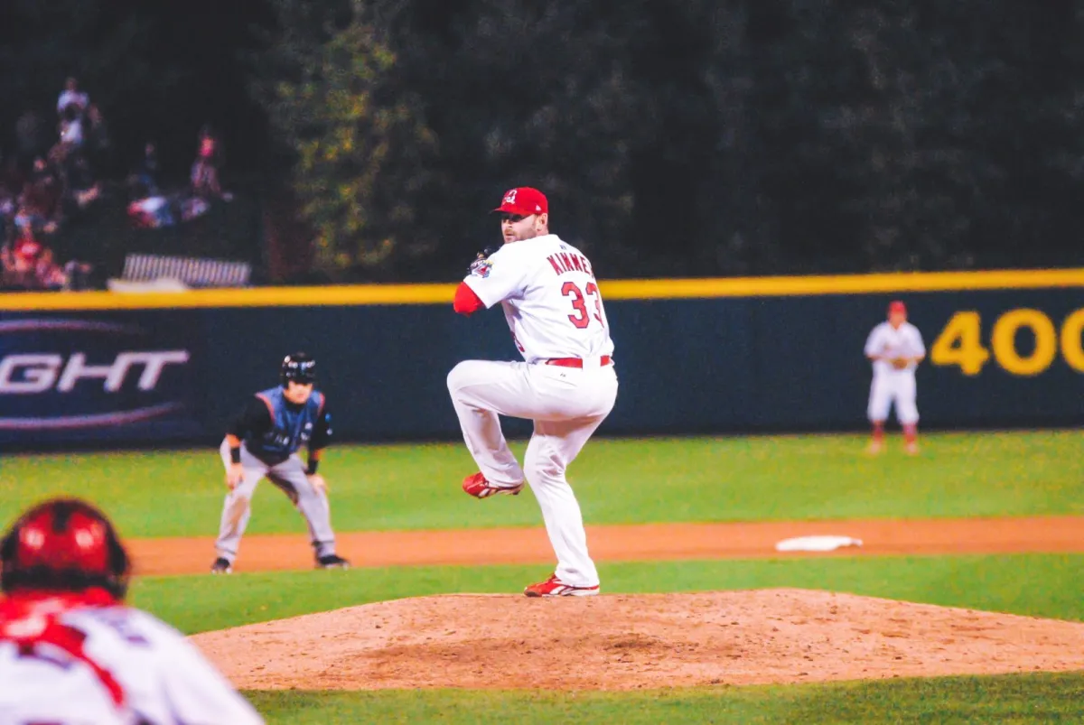 A baseball player in a white uniform and red hat pitches the ball