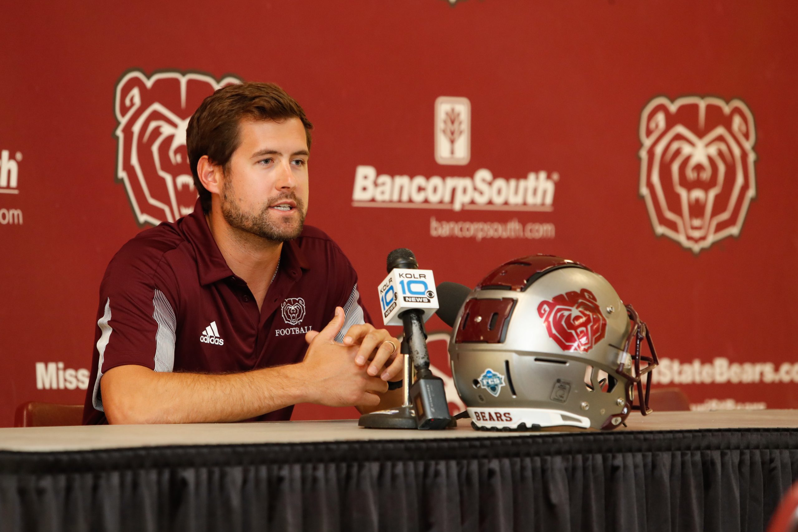 A man in a maroon polo shirt sits at a table and speaks into a microphone