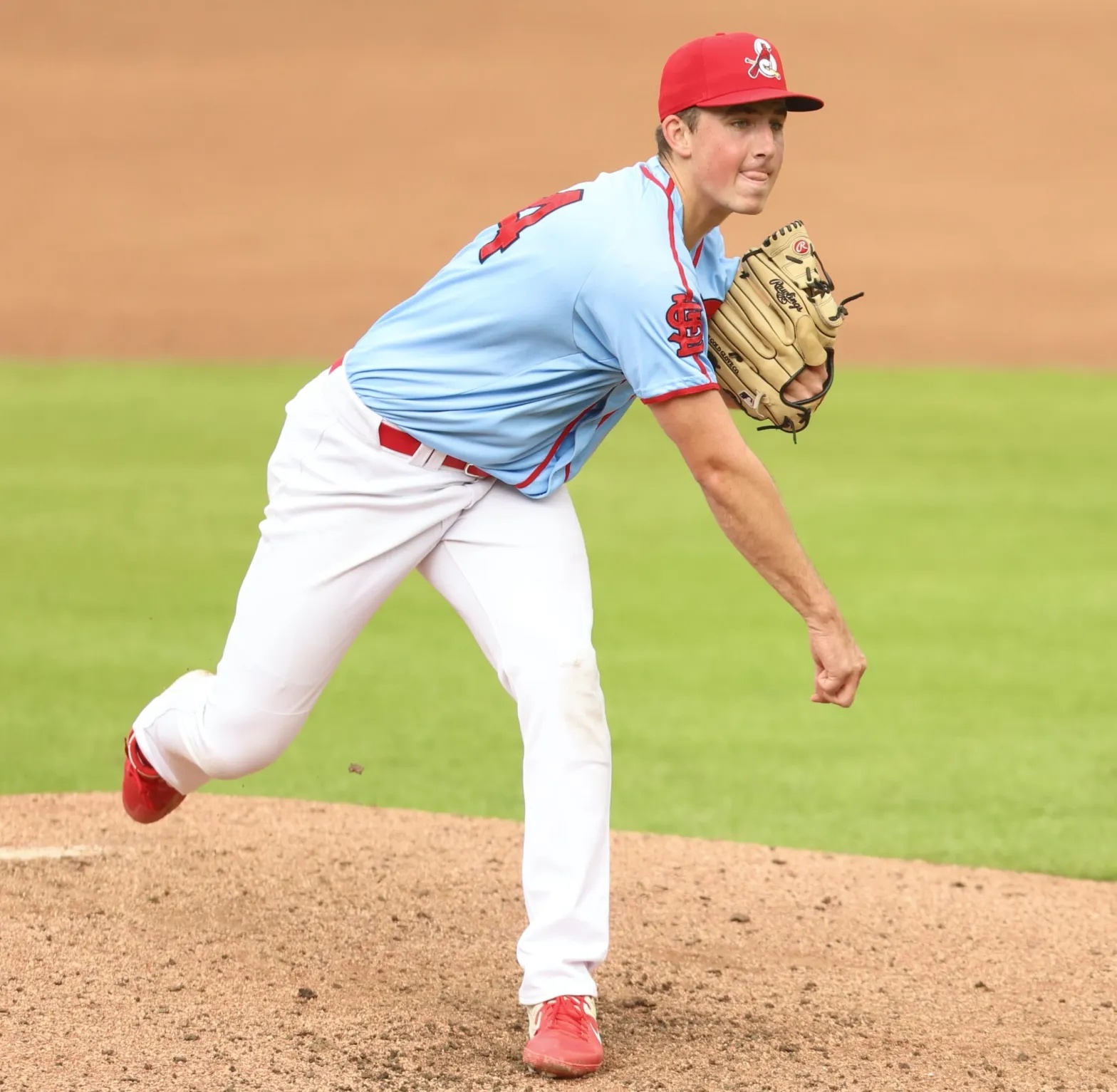 A baseball player in a light blue jersey and red hat pitches the ball