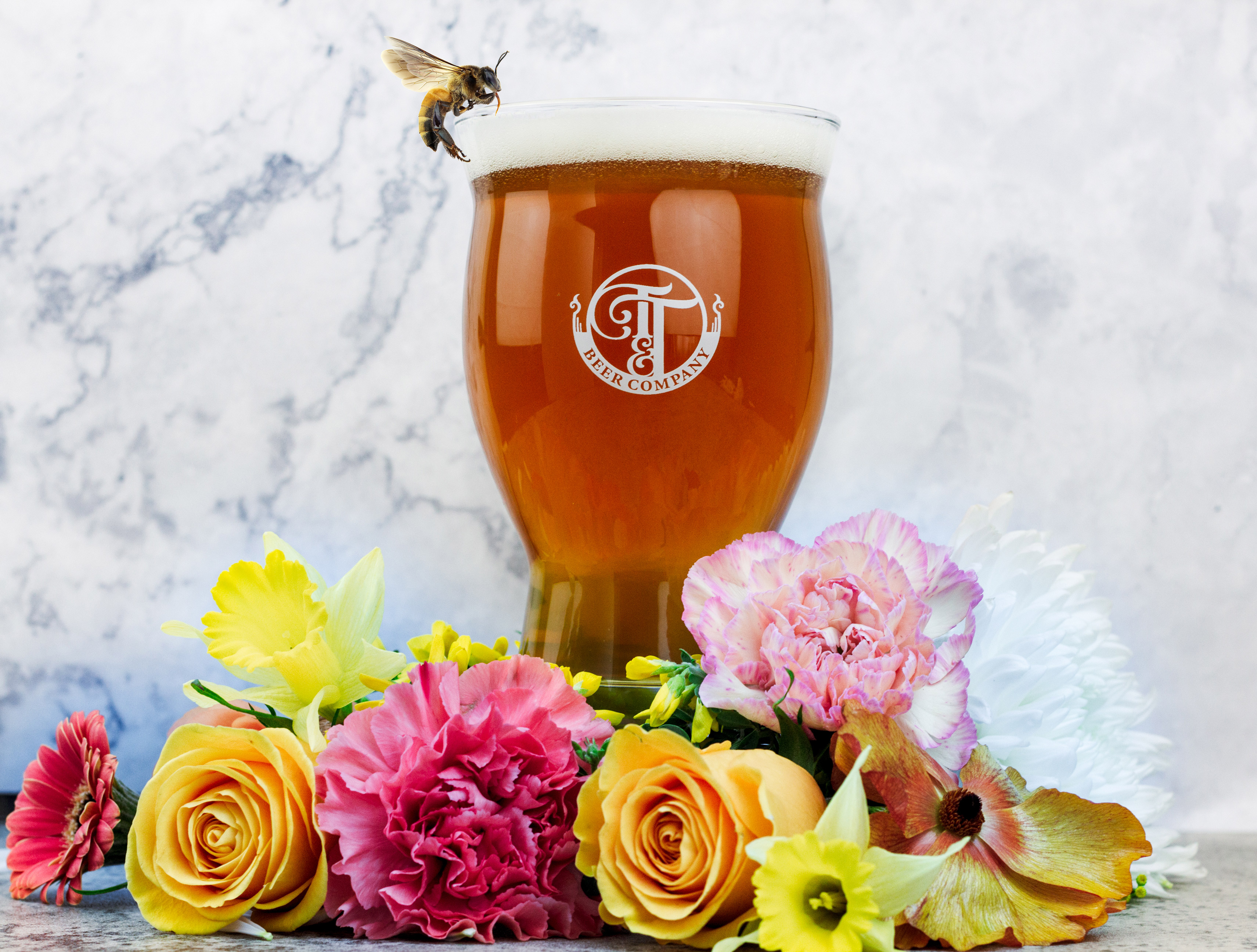 A bee hovers near a glass of beer, with flowers strewn around it