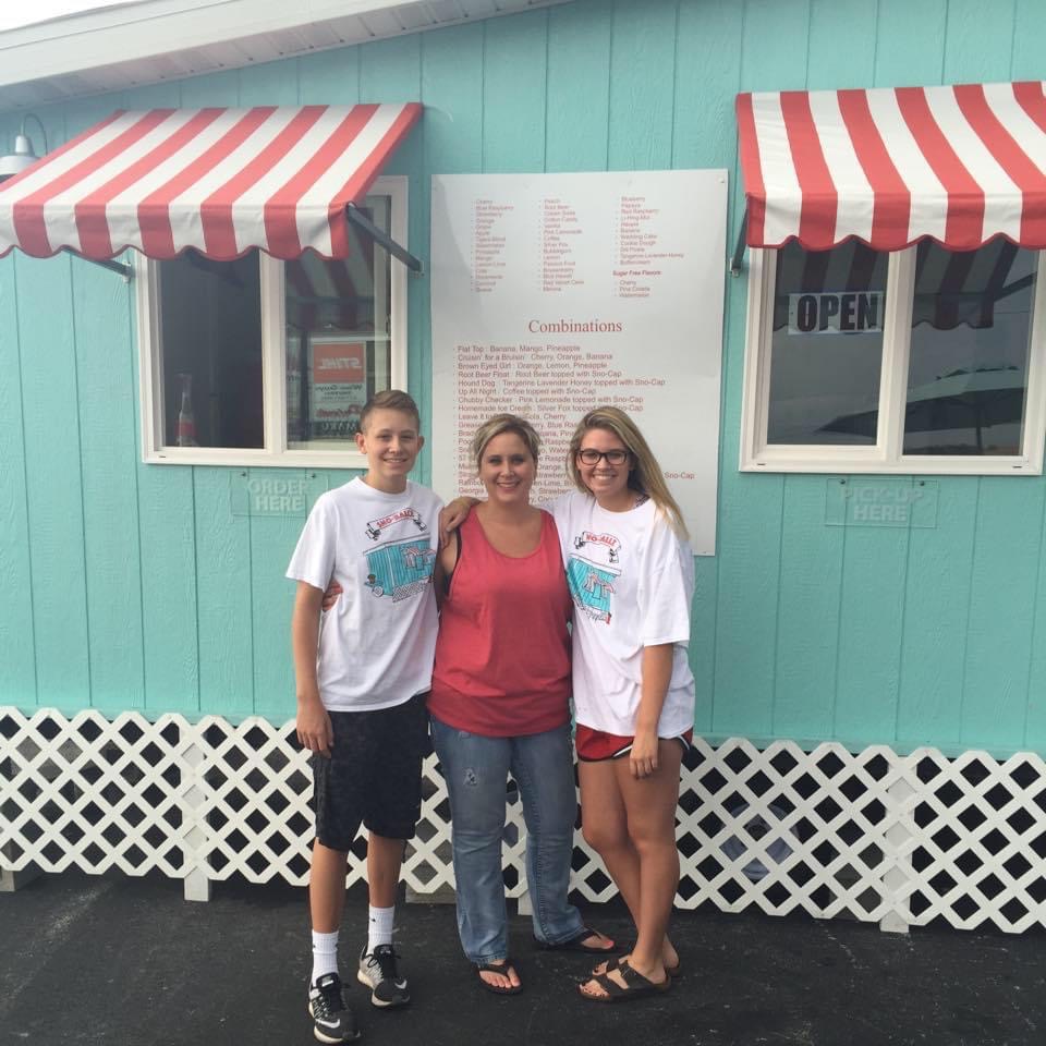 A mother stands between her son and daughter outside a teal building with red and white striped awnings