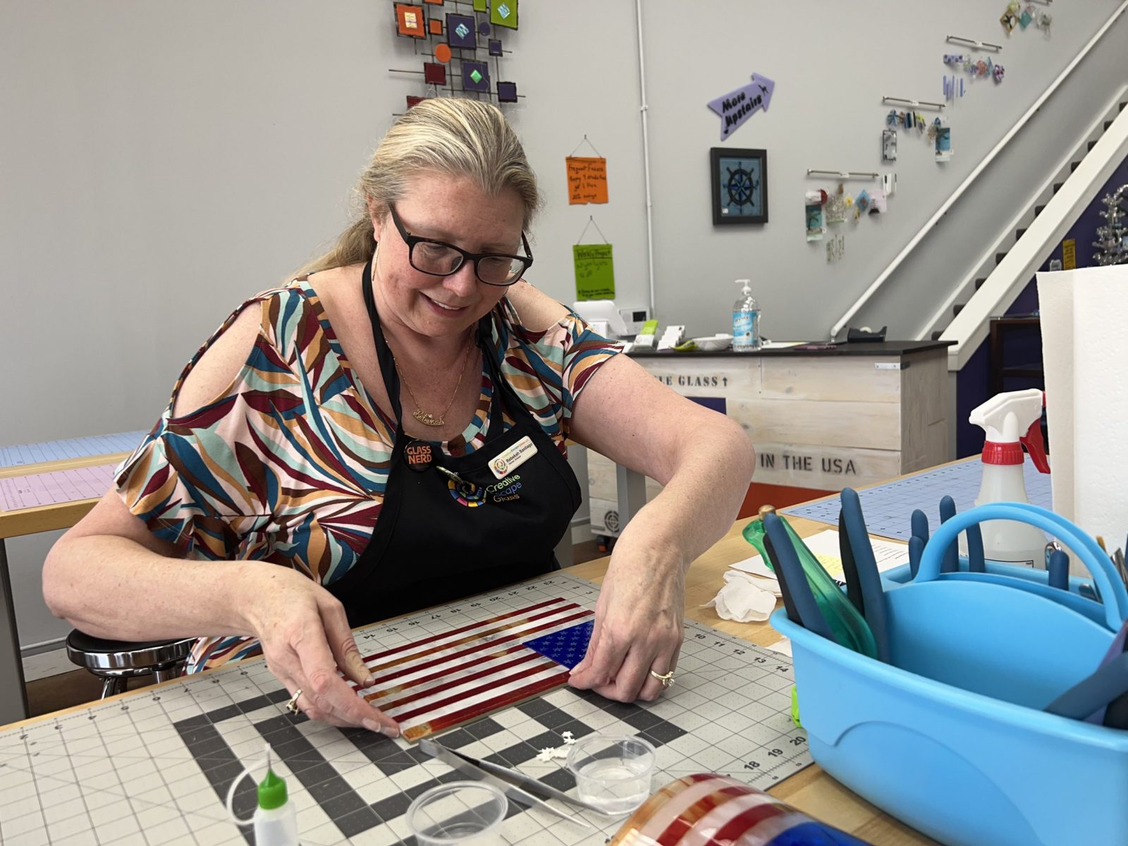 A woman sits at a table, making an American flag from pieces of glass