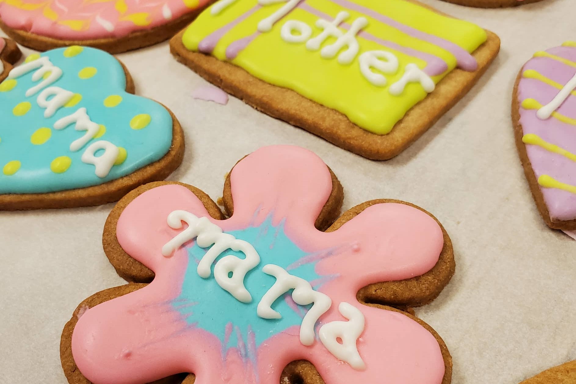 Brightly colored cookies with words like "Mama" and "Mother" written on them in frosting sit on white parchment paper