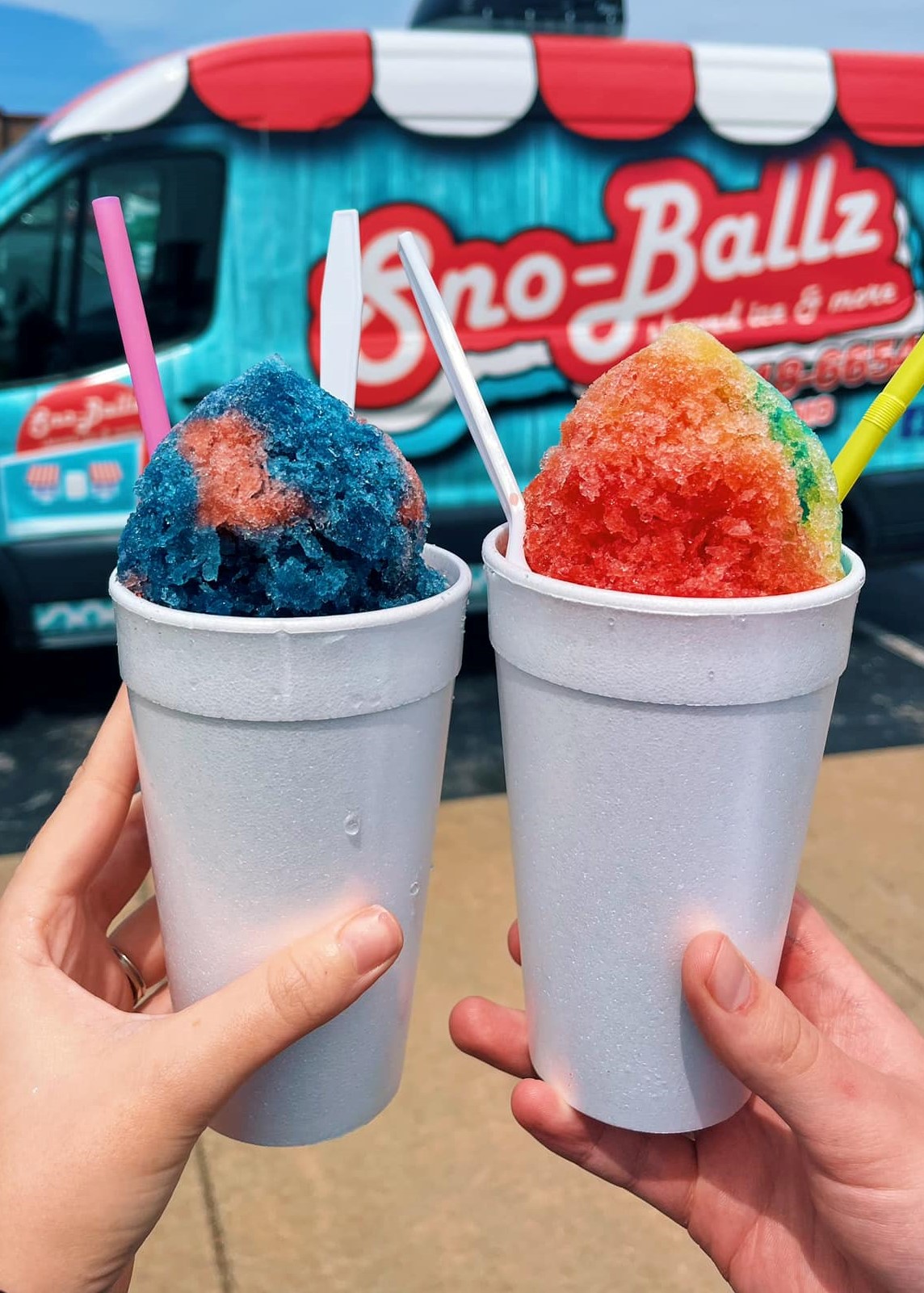 Two hands holing up white styrofoam cups of Hawiian shaved ice in front of a brightly colored food truck