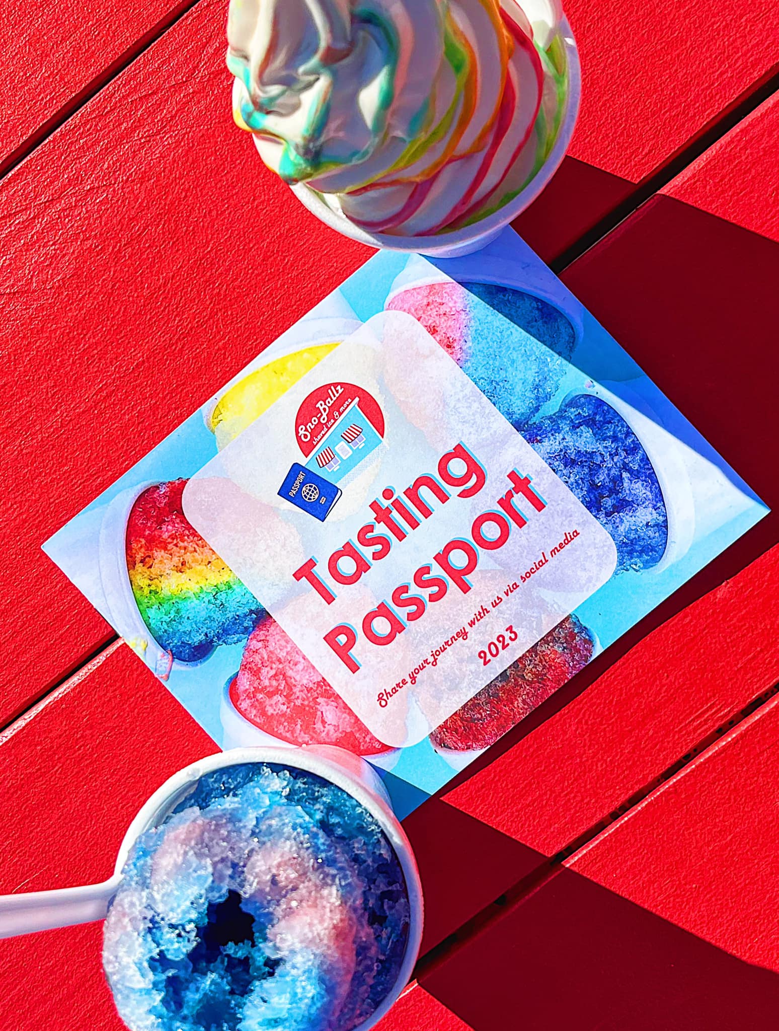A dish of Hawaiian shaved ice and a dish of ice cream sit on a red table next to a "Tasting Passport" from Sno-Ballz