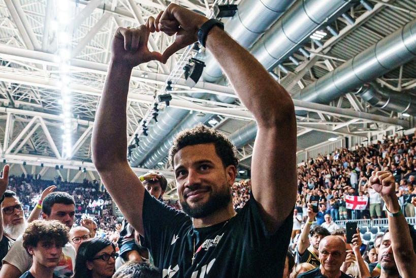 A man in a black T-shirt stands in a crowd, holing up his hands in the shape of a heart