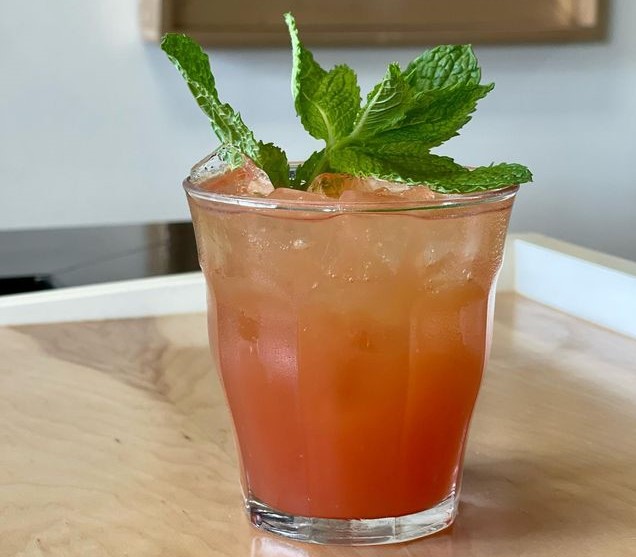 A pink cocktail, garnished with mint, sits on a wooden table