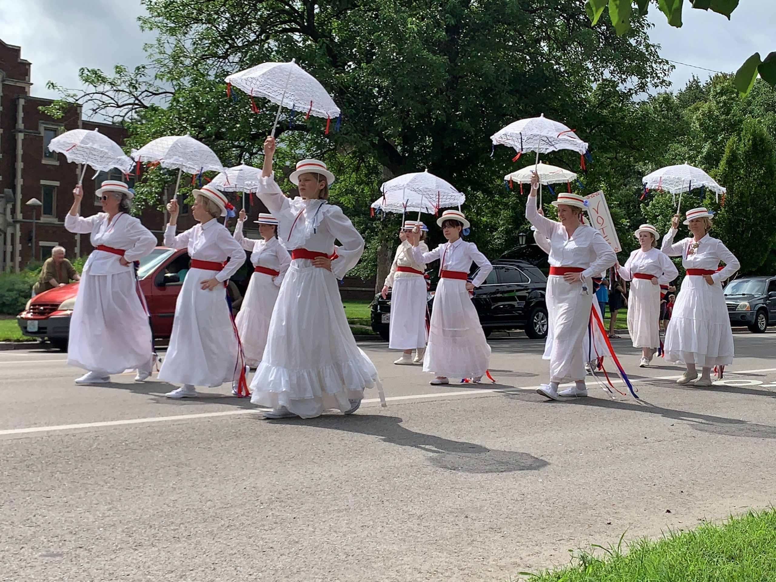A group of women in white dresses holding white umbrellas marches in a parade