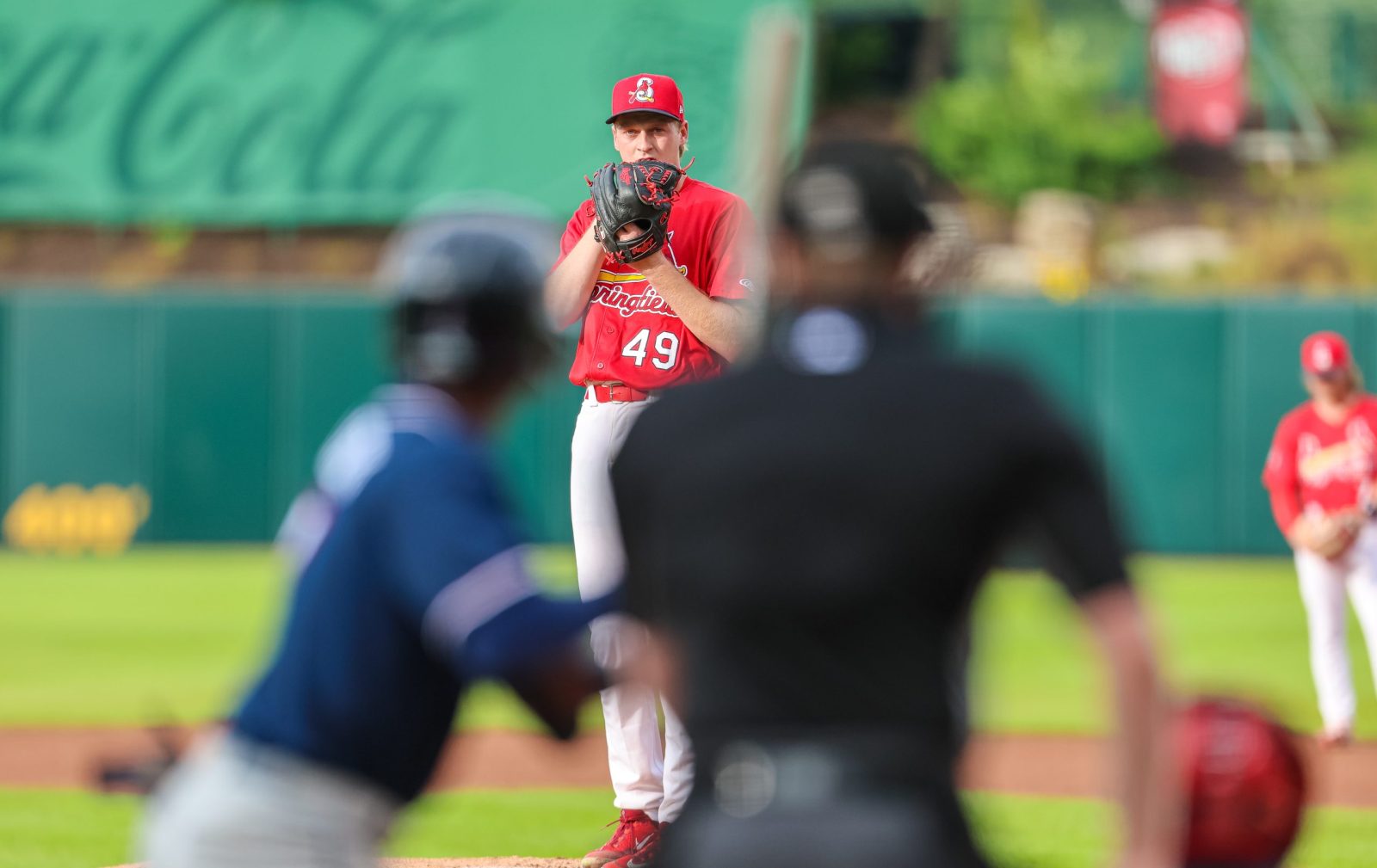 Logan Gragg, wearing a Springfield Cardinals uniform, gets ready to pitch the baseball during a game. He's in the background, with the batter and umpire out of focus in the foreground