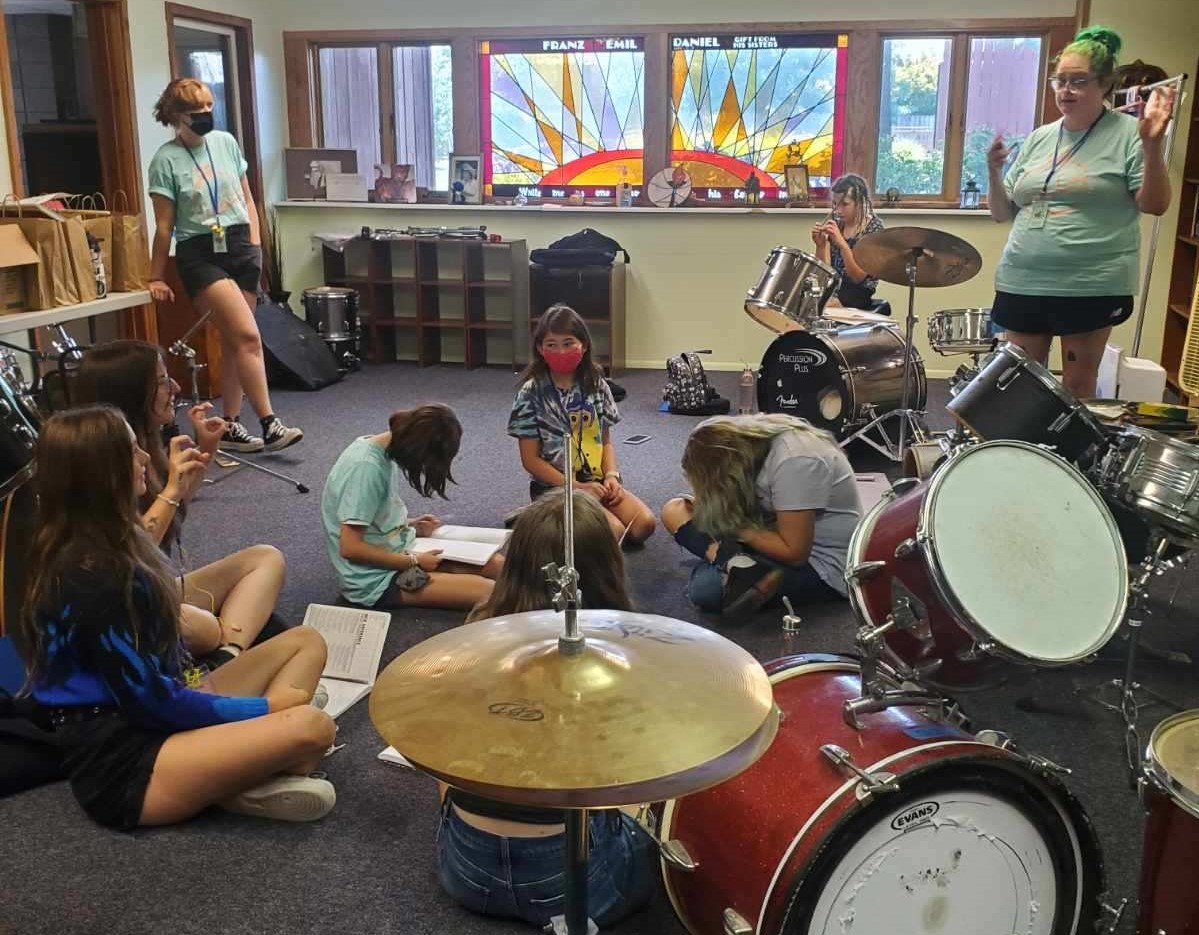 Campers at Queen City Rock Camp sit in a room with a drum kit, participating in a session