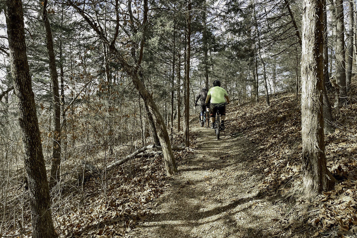 Two cyclists ride on an unpaved trail through a heavily wooded area