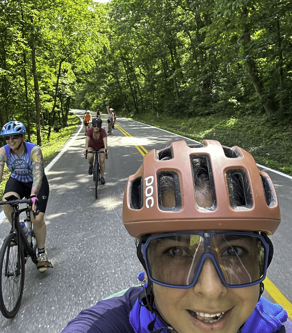 A woman takes a selfie as she rides her bicycle down the road, getting several cyclists behind her in the shot