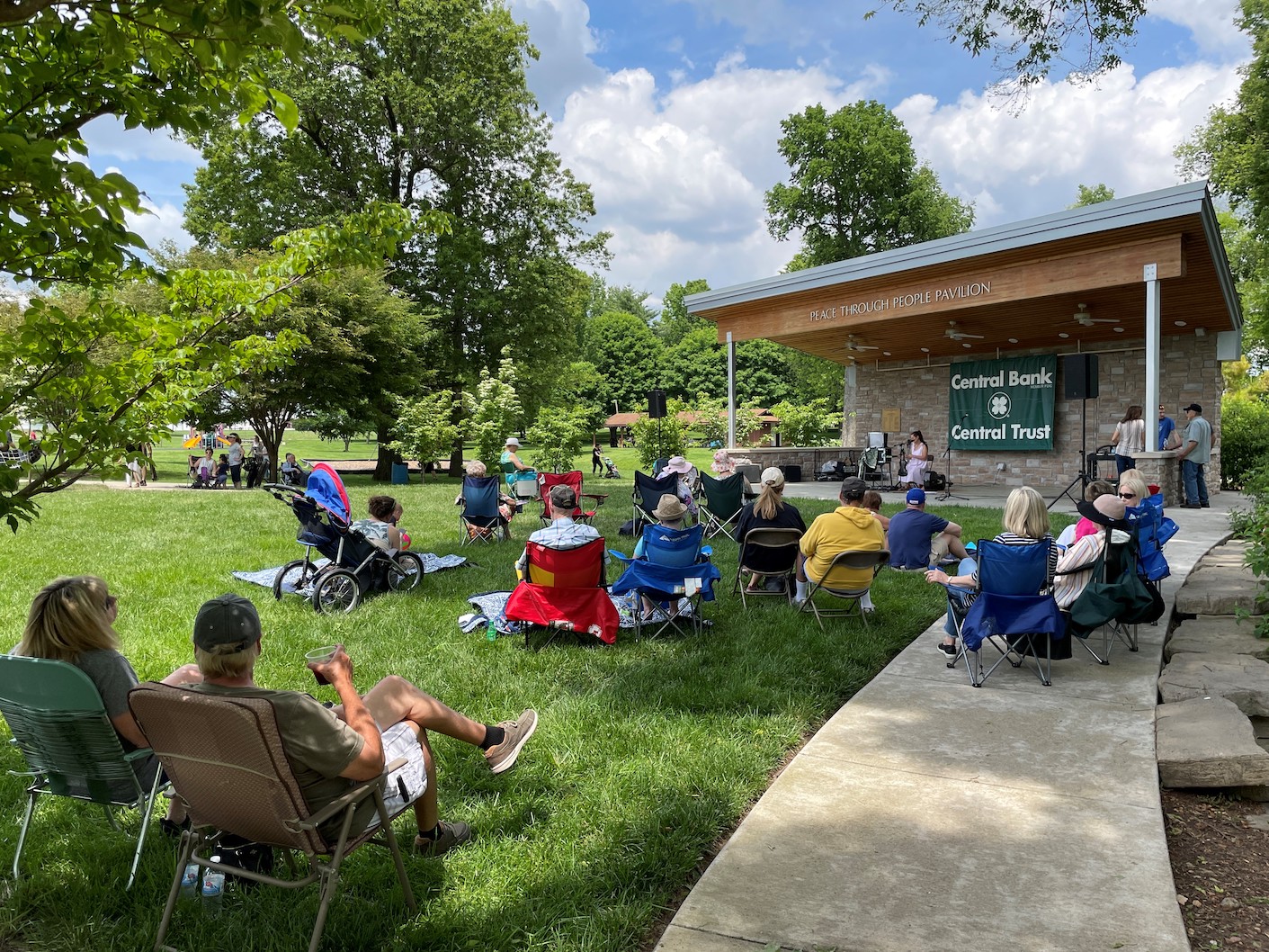 People seated on lawn chairs and blankets enjoy a concert in a park