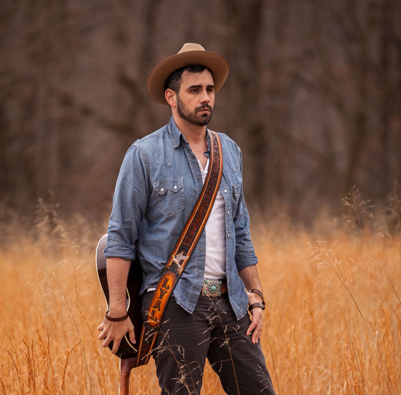 A man in a cowboy hat, wearing a guitar, poses for a photo in a field