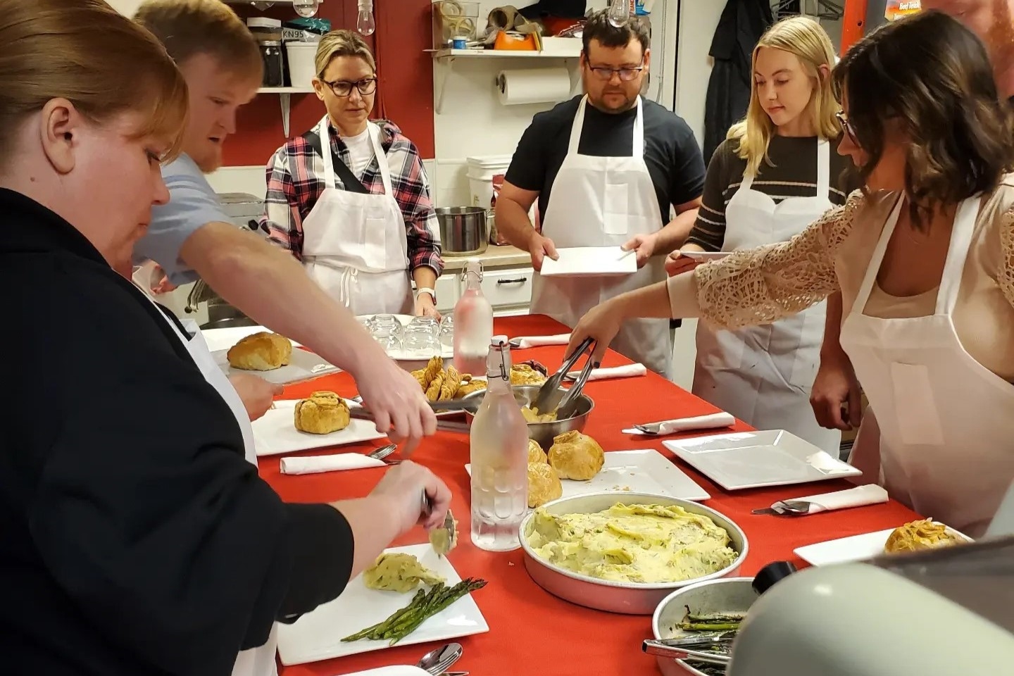 Six people wearing white aprons gather around a red table, taking part in a cooking class