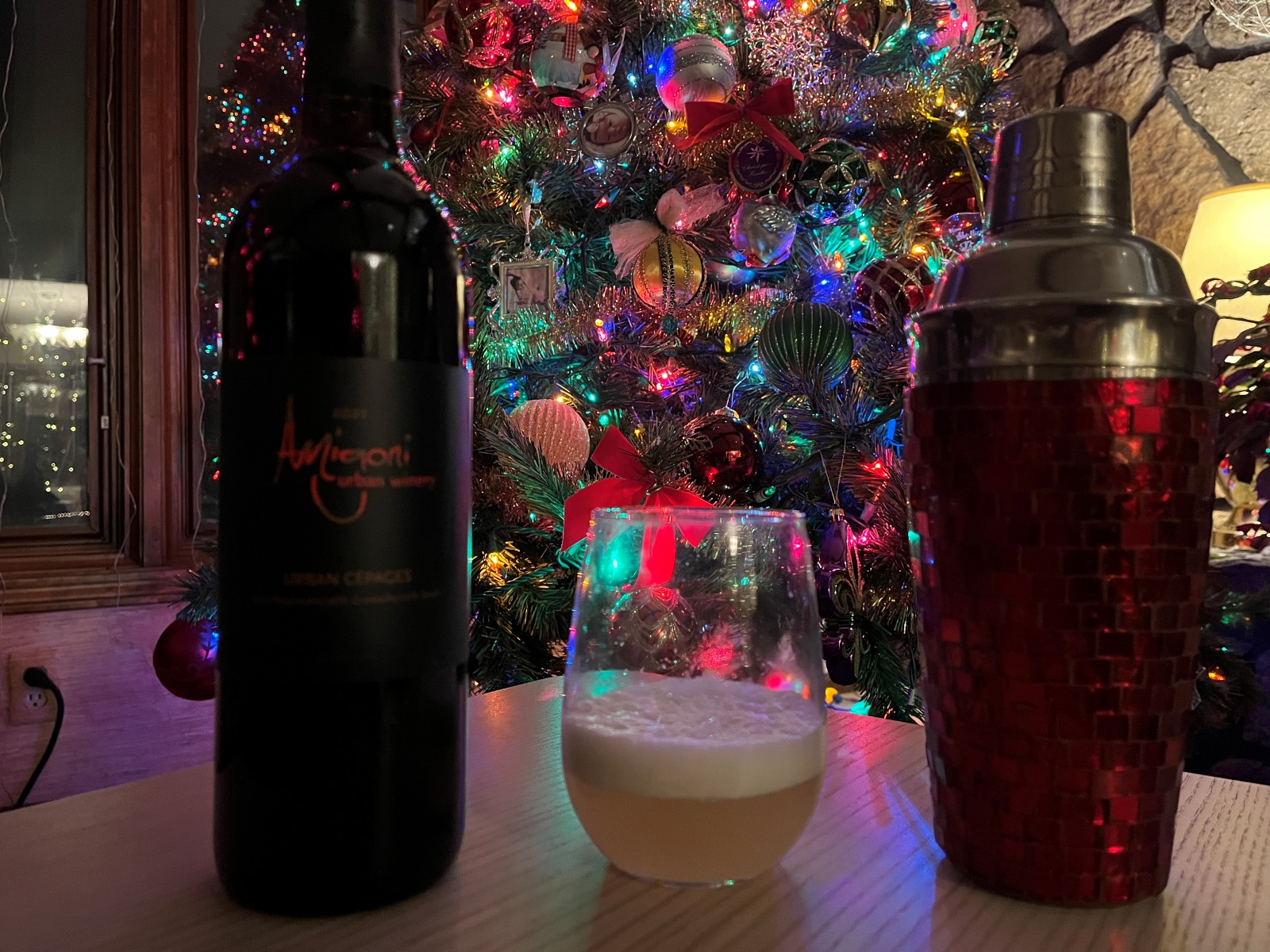 A bottle of wine, a cocktail and a cocktail shaker sit on a table in front of a Christmas tree