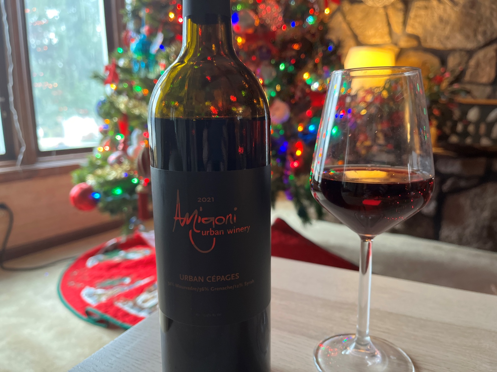 A bottle of Amigoni Urban Winery wine sits on a table, next to a half-full wine glass. In the background is a Christmas tree