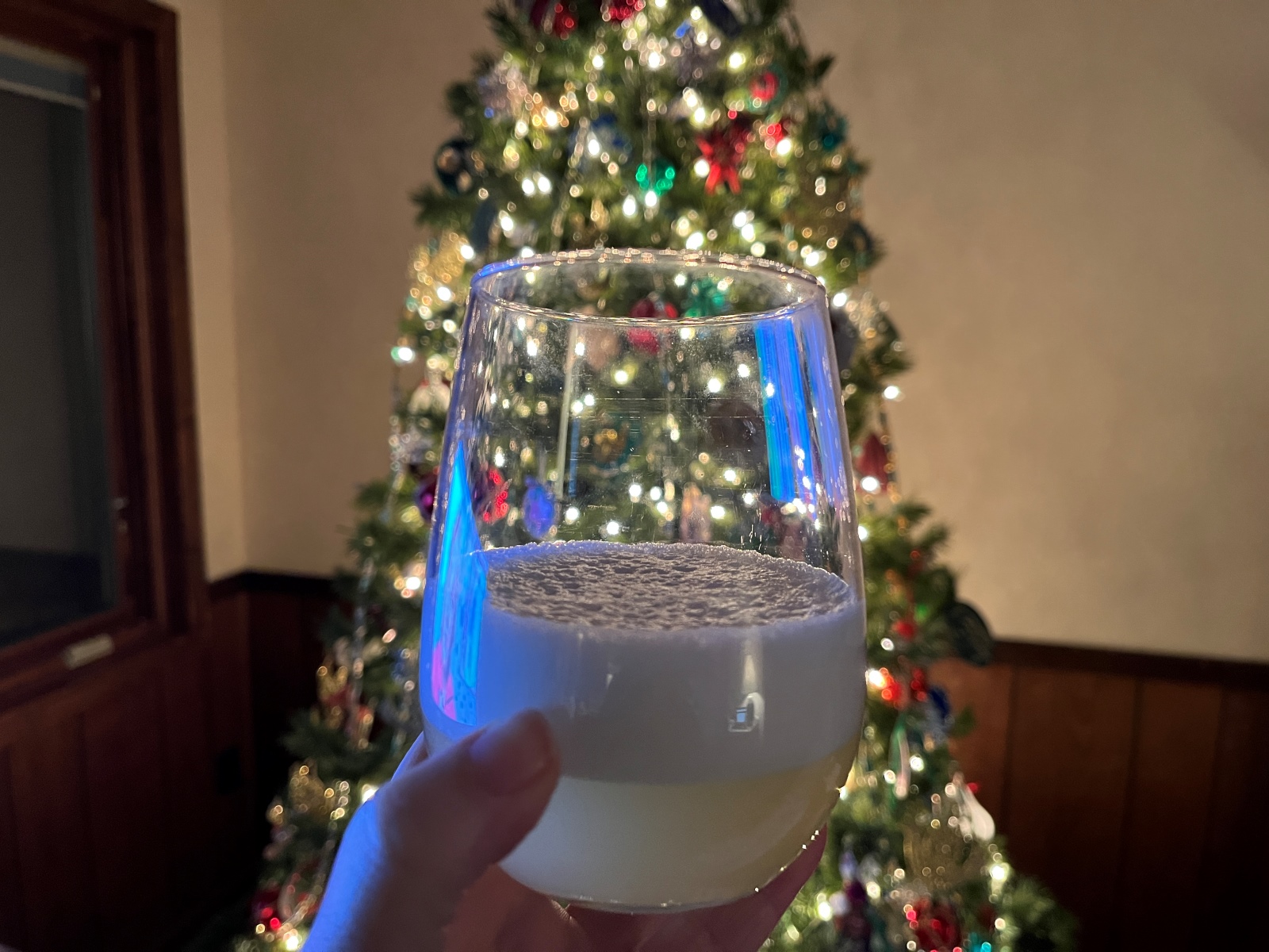 A hand holds a cocktail glass containing a Pisco sour in front of a Christmas tree