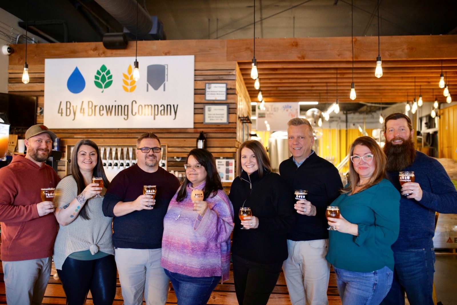 Four couples hold beers and stand in front of the bar at 4 By 4 Brewing Company.