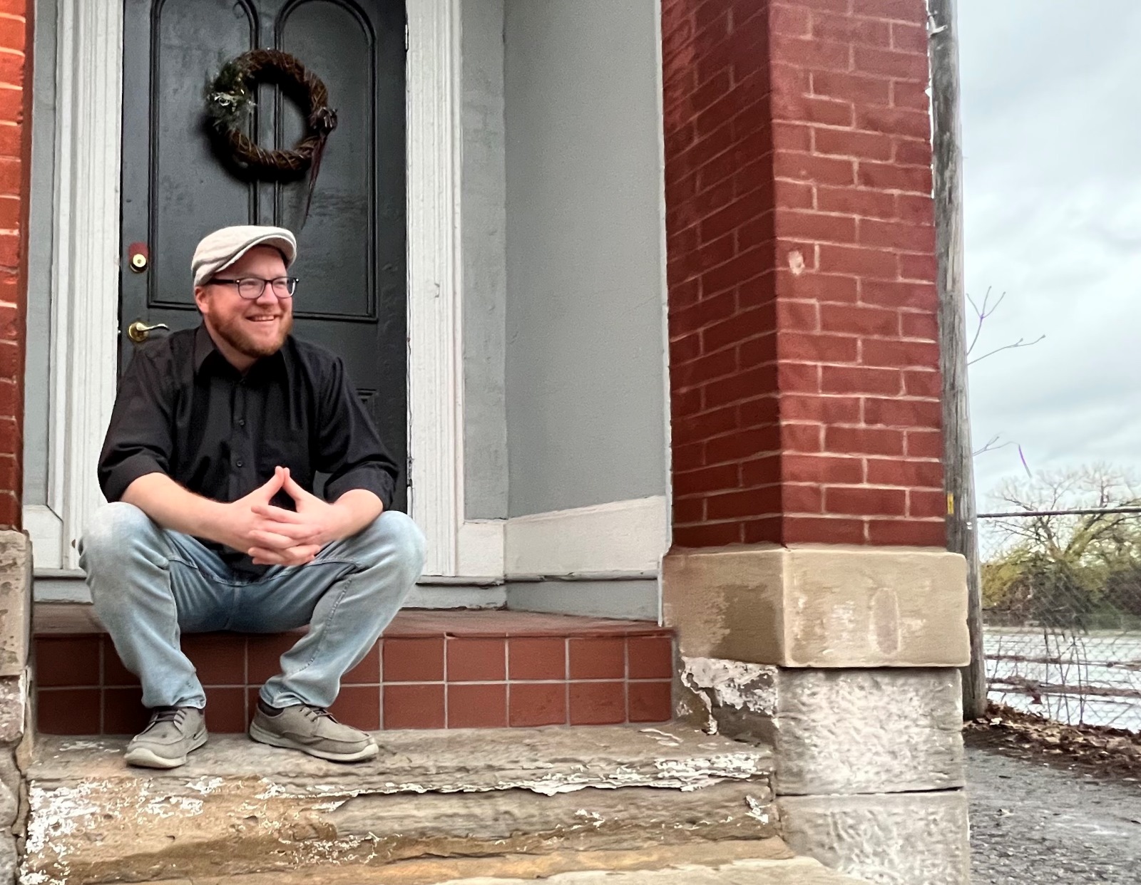 Musician Jimmy Rea, wearing a black button-down shirt and a gray hit, sits on the stoop outside a brick building.