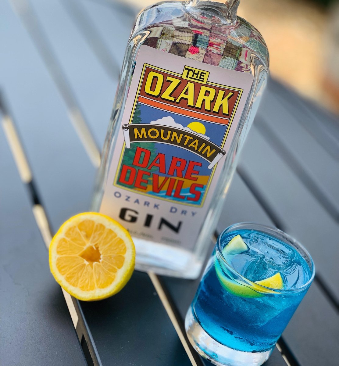 A bottle of The Ozark Mountain Daredevils Ozark Dry Gin sits on a table next to an orange half and a glass with a blue cocktail inside.