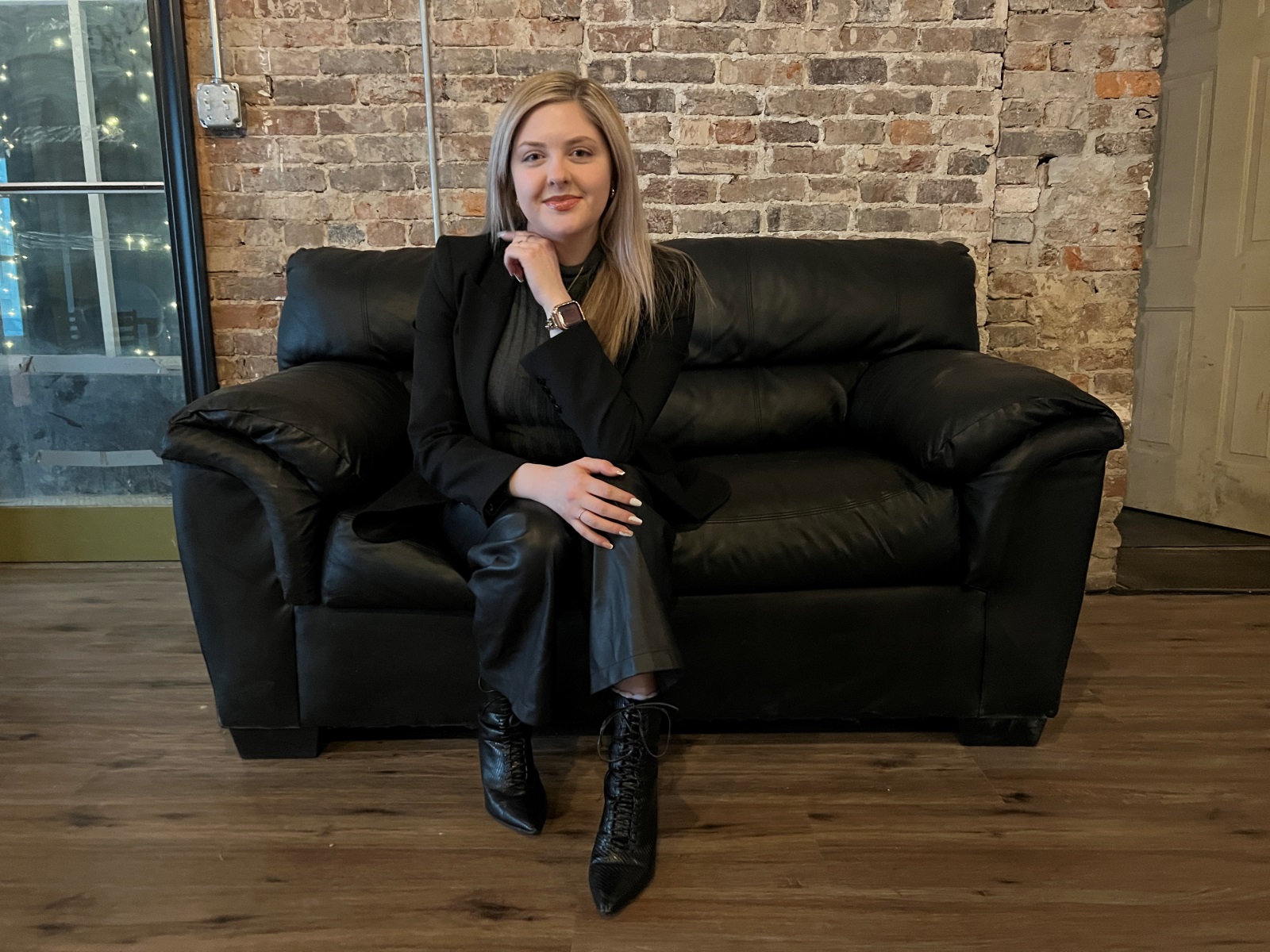 Grace Billingsley, wearing all black, sits on a black couch in front of a brick wall