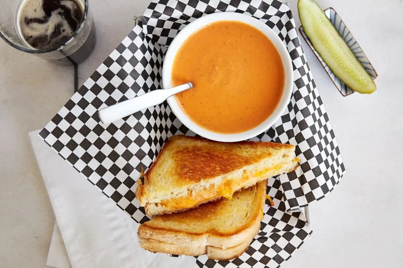 A grilled cheese sandwich sits on a platter next to a bowl of tomato soup and a pickle.