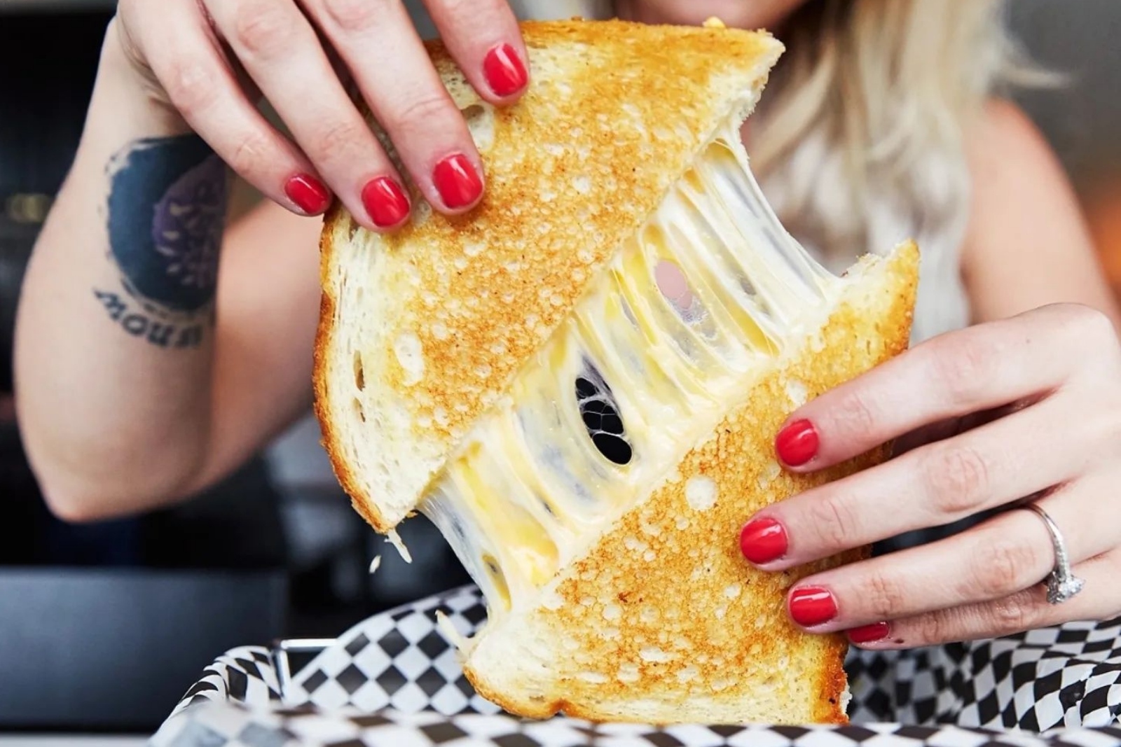 A woman sitting at a table in a restaurant holds a grilled cheese sandwich as the cheese between the slices stretches