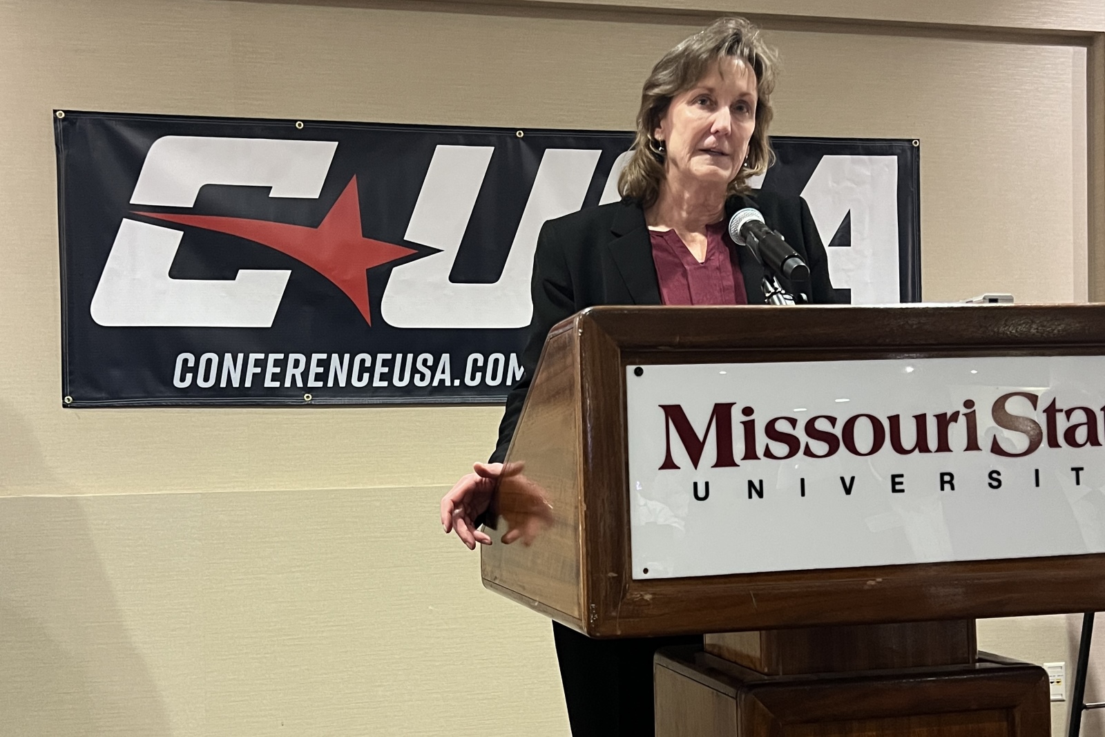 Judy MacLeod, commissioner of Conference USA, stands behind a podium with a sign reading "Missouri State University" and in front of a blue banner with the Conference USA logo printed on it.