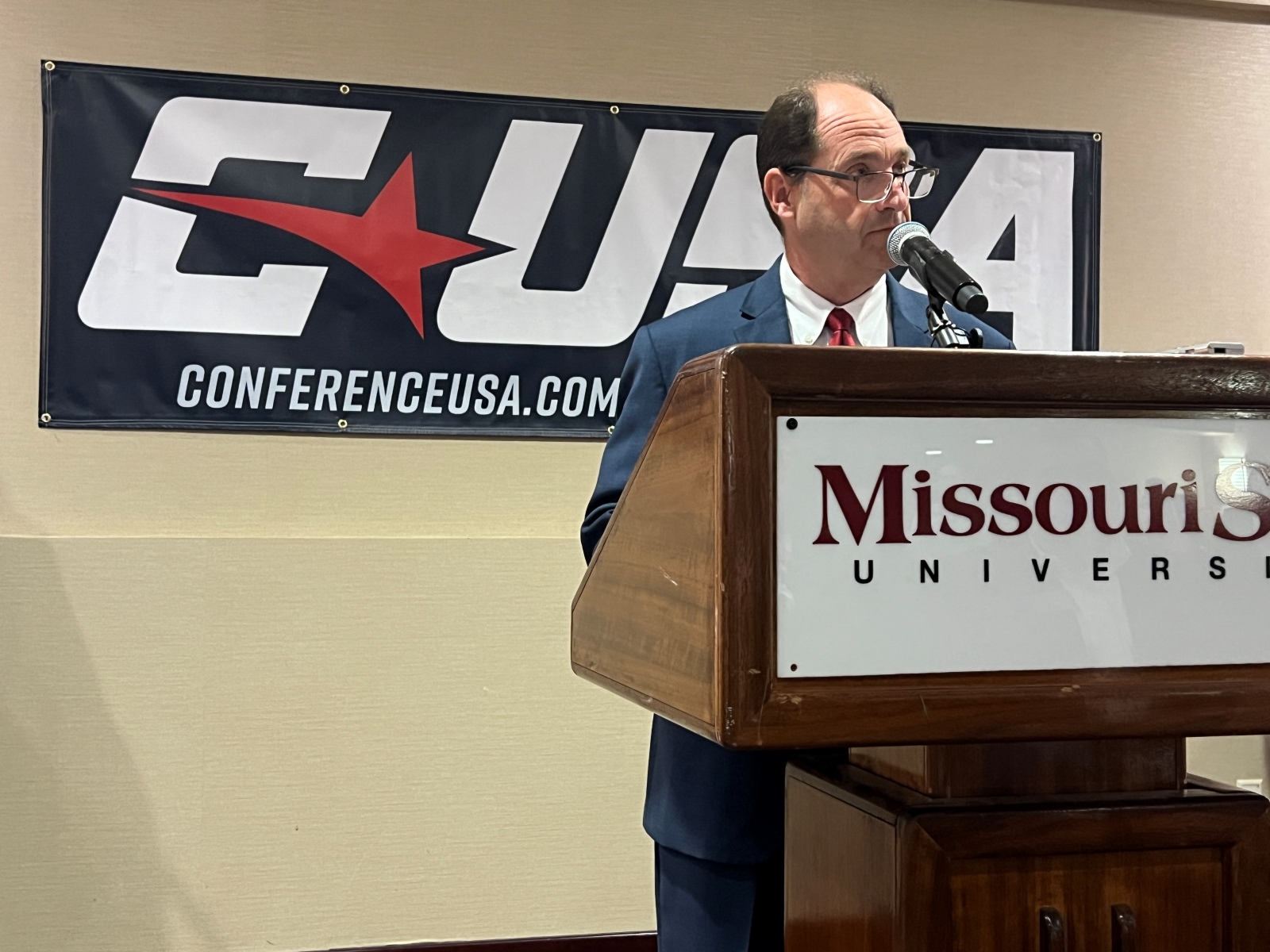 Missouri State athletics director Kyle Moats stands behind a podium with a sign reading "Missouri State University" and in front of a blue banner with the Conference USA logo printed on it.