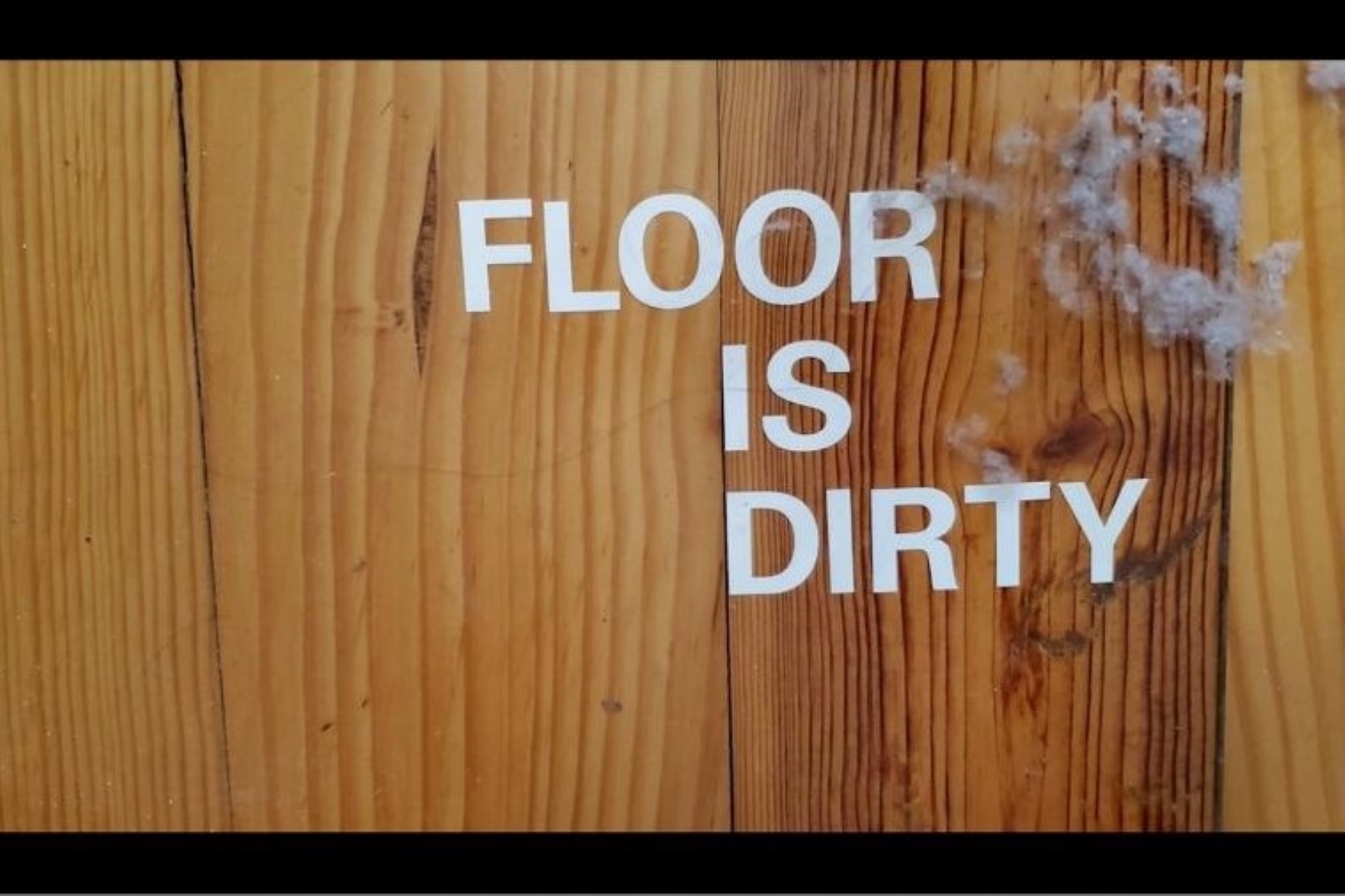 A wood floor with a dust bunny on it. White letters on the floor read "Floor is dirty."